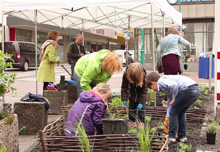 Erwachsene und Kinder legen in einer Fußgängerzone Beete an, die mit Flechtzäunen umgrenzt sind