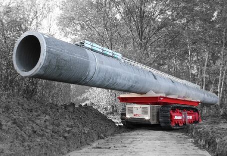 ein kleines selbstfahrendes Raupenfahrzeug transportiert auf seiner Ladefläche einen großen Betonmast durch den Wald