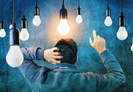 Ein Mann steht mit dem Rücken zum Betrachtenden. Kratzt sich am Kopf und zeigt mit der rechten Hand in die Luft. Über ihm hängen viele Glühbirnen, von denen eineüber seinem Kopf leuchtet.