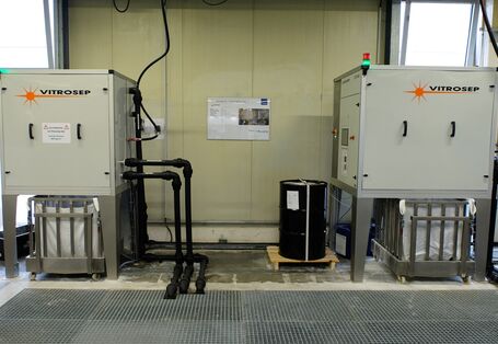 teschnische Anlage in einer Werkshalle aus zwei Boxen, aus denen Rohre in eine Gitterabdeckung im Fußboden führen