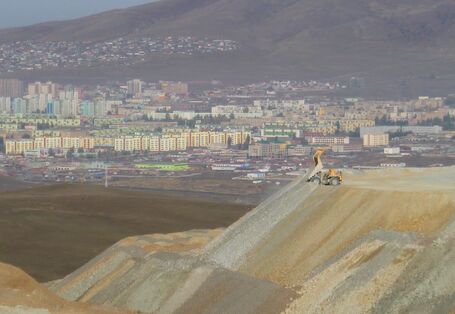 riesige, terrassierte Halde aus Gestein vor einer am Berg gelegenen Stadt mit Hochhaussiedlungen. Ein Muldenkipper kippt weiteres Material ab.