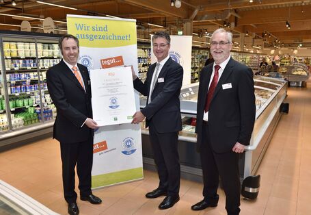 drei Männer in Anzügen stehen nebeneinander in einem Supermarkt und lächeln in die Kamera, zwei halten gemeinsam eine Urkunde mit dem "Blauen Engel" darauf , im Hintergrund ein Plakat mit dem "Blauen Engel" und der Aufschrift "Wir sind ausgezeichnet!"