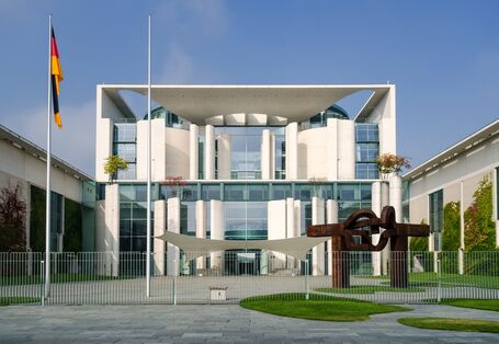 modernes weißes Gebäude mit Flachdach und großzügigem Eingangsbereich mit verglaster Fassade, davor eine deutsche Flagge und eine moderne Gartengestaltung mit amorphen kleinen Rasenflächen und modernen Skulpturen
