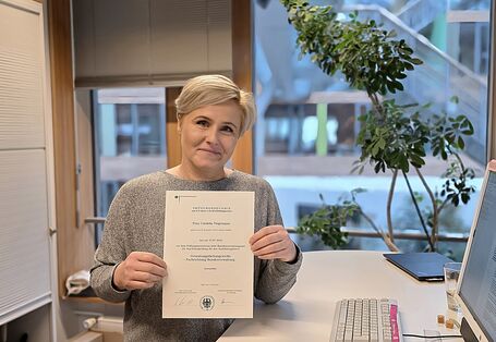 Cordelia Feigenspan hält in ihrem Büro neben ihrem Schreibtisch stehend ihr Prüfungszeugnis in die Kamera