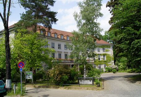 Three-storey historic building with entrance portal, with balcony and flower boxes above, gabled roof with many small dormers. At the front a square with trees, bushes, a drive and sign which reads "Umweltbundesamt"