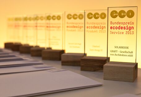 Auf einem Tisch stehen in einer Reihe die Sieger-Trophäen aus Glas mit dem Logo des Bundespreis Ecodesign und dem Namen des jeweiligen Preisträgers