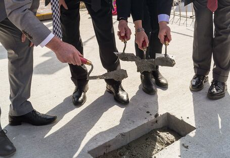 drei Männer und eine Frau in festlicher Kleidung betonieren mit Maurerkellen eine quadratische Aussparung in einer Bodenplatte zu und lächeln in die Kamera