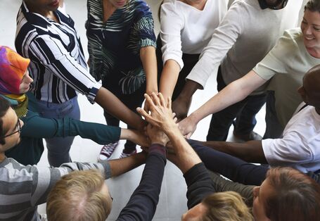 Frauen und Männer mit verschiedenen Hautfarben, mit und ohne Kopftuch stehen in einem Kreis und halten ihre Hände gemeinsam in die Mitte aufeinander