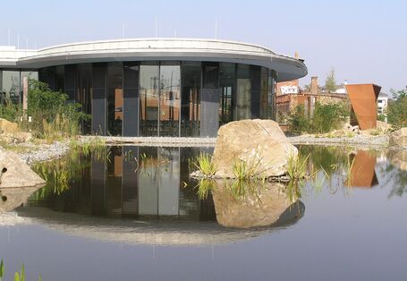 Low, round building with glass façade reflected in a pond surrounded by natural stones
