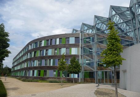 Haupteingang des UBA Dessau-Roßlau mit Glasfassade, die restliche Fassade besteht aus Holz, Fenstern und Glasplatten in Grün- und Blautönen
