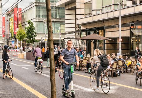 Fußgängerzone mit Radspur, Tischen vor Cafés, Geschäften und Bäumen in Kübeln mit Sitzgelegenheiten
