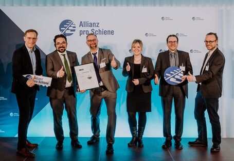 fünf Männer im Anzug und eine Frau im Kostüm stehen lächelnd mit einer Urkunde für ein Gruppenfoto postiert vor einer Stellwand mit dem Logo "Allianz pro Schiene"