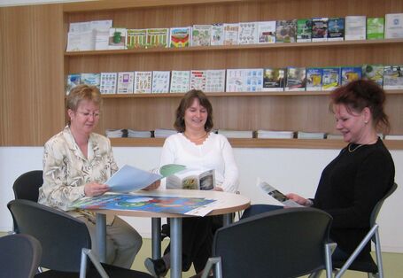 Three women sitting at a round table looking at flyers and brochures, a shelf with more materials in the background