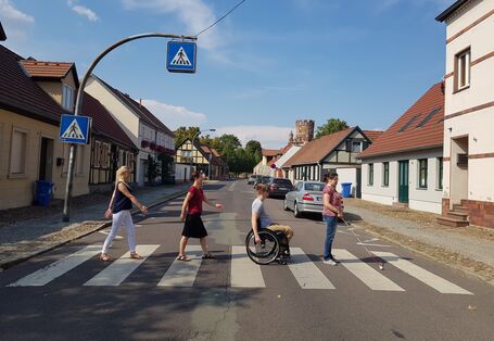 Straße mit niedrigen Fachwerkhäusern und einem Zebrastreifen, der von einer Frau mit Blindenstock, einem Rollstuhlfahrer und zwei weiteren Fußgängerinnen überquert wird.