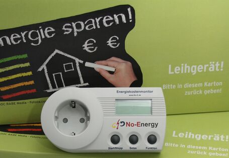 Das Bild zeigt eine hellgrüne Pappkiste mit einem Messgerät für Leerlaufverluste von Elektrogeräten im Vordergrund
