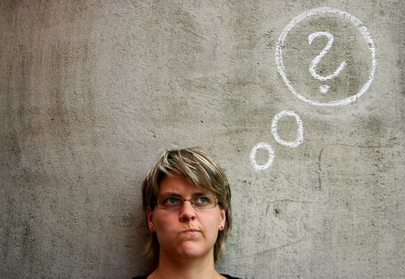 Eine Frau mir fragendem Blick steht vor einer Wand, auf die mit Kreide ein Fragezeichen gemalt ist.