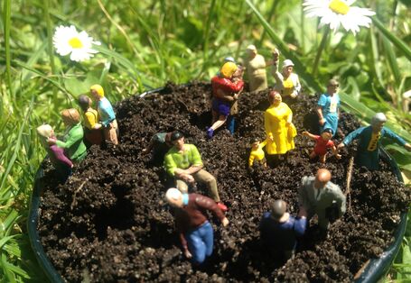 Ein Eimer voller Erde steht auf einer Wiese, darin stecken mehrere Spielzeugfiguren.