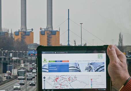 Vor einem Energiekraftwerk hält eine Hand einen Tablet-PC hoch. Auf dem Display sind Informationen zu diesem Kraftwerk zu sehen.