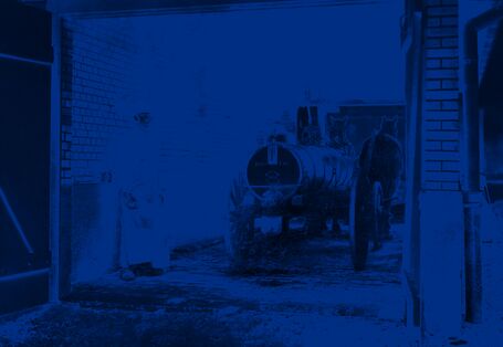 Ein Mann steht mit einem Jauchewagen in einer Einfahrt, der Wagen trägt einen Metalltank und wird von zwei Pferden gezogen. 