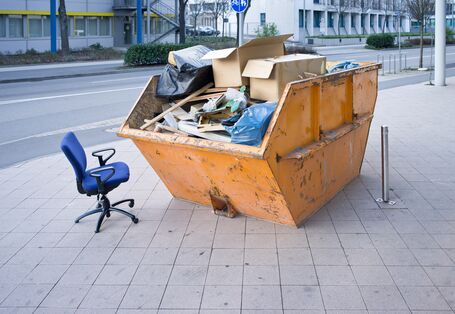 Ein Müllcontainer steht auf der Straße, daneben steht ein Bürostuhl