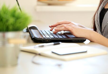 Eine Frau schreibt auf einem Notebook. Vor ihr steht ein Blumentopf mit einer Grünpflanze.