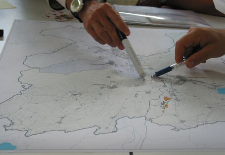 Nahaufnahme von zwei Händen unterschiedlicher Personen, die auf einem Plan mit Stiften auf einen gemeinsamen Ort zeigen.