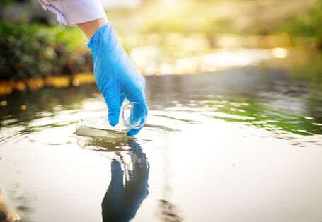Foto Hand mit Plastikhandschuh nimmt Wasserprobe in Glasgefäß aus einem Gewässer.