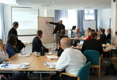 Man sieht ein Bild von dem Stakeholderdialog 2016: Indirekte Effekte des globalen Klimawandels auf die deutsche Wirtschaft. Im Hintergrund sieht man eine Präsentation und eine Person, die auf die Folien zeigt. Drumherum sitzen mehrere Personen an Gruppentischen und schauen gespannt nach vorne. 