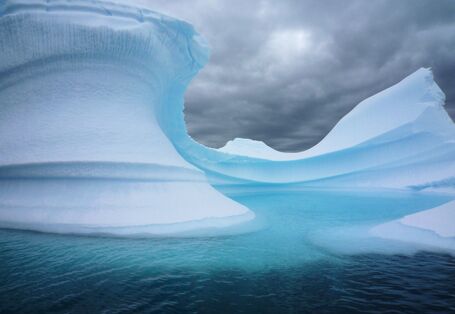Vor dunkel bewölktem Himmel ragt ein weiß-blauer Eisberg auf. Er ist geschwungen. Das Wasser um ihn ist dunkelblau.
