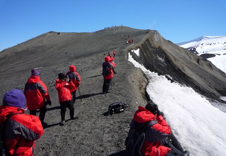 Touristen in roten Funktionsjacken erklimmen einen Hügelgrat. Der Hügel ist nur zur Hälfte schneebedeckt. Einige Leute machen Photos.