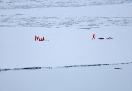 Auf einer weiten Eisebene steht eine Gruppe von Forschern und roten Kälteschutzanzügen. Sie entnehmen an einer Stelle eine Eisprobe. Weiter rechts läuft eine weitere Person zur Ausrüstung. 