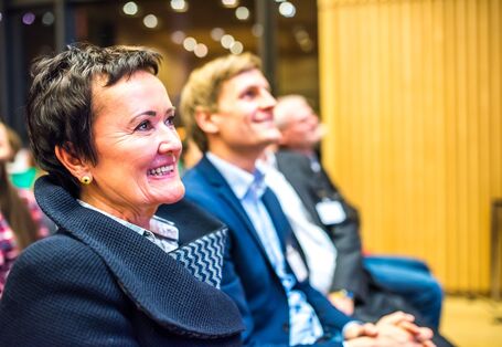 Es ist das Profil einer Frau zu sehen. Sie schaut nach vorne und lacht. Sitzt mit anderen Leuten in einer Stuhlreihe im Publikum und auch der Mann links neben ihr grinst interessiert.