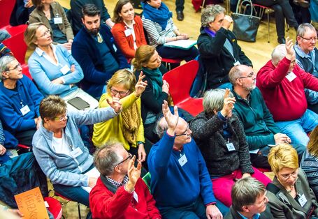 Es sind viele Menschen in 5 Stuhlreihen zu sehen. Sie sind von oben fotografiert. Es ist ein fröhliches Bild, die Menschen wirken sehr aufgeweckt und tragen bunte Kleidung. Die Menschen melden sich und beteiligen sich rege.