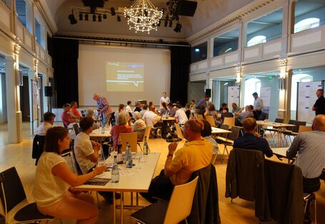 Die Teilnehmenden sitzen in einem großen Saal mit Bühne und schauen auf die vorgetragenen Präsentationsfolien.
