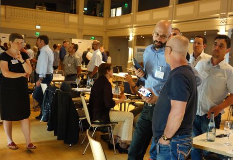 Die Teilnehmenden stehen in einem großen Saal neben Sitzgruppen mit Stühlen und Tischen. Sie tauschen sich aus und mischen sich untereinander durch.