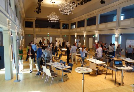 In einem prunkvollen Saal mit Kronleuchter sind einige Sitzgruppen mit Tischen und Stühlen zu sehen. Um diese stehen oder bewegen sich die Teilnehmenden herum. Sie bilden Grüppchen und tauschen sich aus.