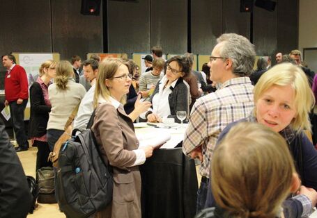 Die Teilnehmenden stehen verteilt um Stehtische und tauschen sich angeregt aus. Im Vordergrund steht eine blonde Frau und in der Mitte eine Frau mit einem schwarzen Rucksack, die mit einem Herren im karierten Hemd spricht. Hinter ihnen sind noch sehr viele weitere Personen zu sehen.