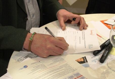 Auf dem Bild ist ein Stehtisch zu sehen in der Draufsicht und zwei Hände. Auf dem Tischen befinden sich mehrere Blätter und ein Glas. Eine Person füllt ein Blatt mit der rechten Hand aus. Die Person trägt ein buntes Armband.