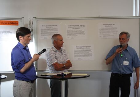 Vor einer weißen Tafel mit aufgeklebten Blättern mit Texten stehen drei Männer. Zwei der drei Männer halten ein Mikrofon. Der eine spricht in dieses hinein. In der Mitte ist ein Stehtisch.