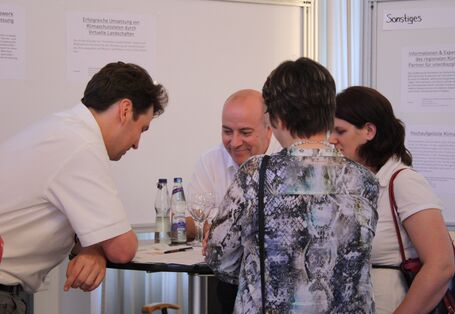 Um einen Stehtisch herum stehen vier Personen. Auf dem Tisch befinden sich Gläser und Wasser sowie Zettel und Stifte. Alle Personen schauen auf den Tisch und lesen.