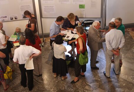 Das Foto wurde von oben aufgenommen und Personen sind von oben zu sehen. Diese stehen um zwei Stehtische herum und neben diesen. Einige lesen, andere reden miteinander. Es ist auch ein kleines Kind unter ihnen.