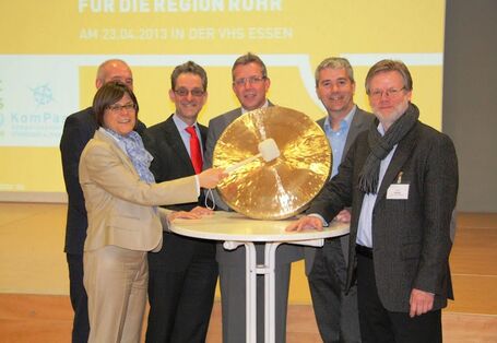 Hinter einem Stehtisch stehen sechs Personen. Sie posieren für ein Foto. Eine Frau hält eine große goldene Klangplatte und einen hölzernen Hammer,  mit dem sie auf die Platte schlägt, um einen Gong zu erzeugen. 
