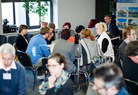 Die Teilnehmenden sitzen in Stühlkreisen und Tischen und tauschen sich angeregt aus. Im Vordergrund sind drei Personen zu sehen. Im Hintergrund sitzen 8 Personen in einem Kreis.