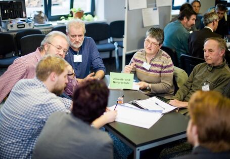 Sieben Personen sitzen um einen schwarzen Tisch. Sie tragen Namensschilder und diskutieren angeregt. Auf dem Tisch liegen verschieden Zettel. Eine Person füllt ein Blatt aus.
