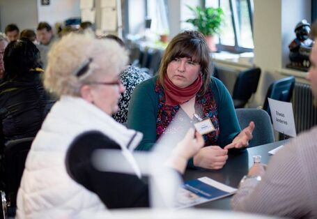 Eine ältere Dame mit lockigen weißen Haaren sitzt neben einer Frau mit Ponyfrisur an einem Tisch. Sie reden miteinander und gestikulieren mit den Händen.