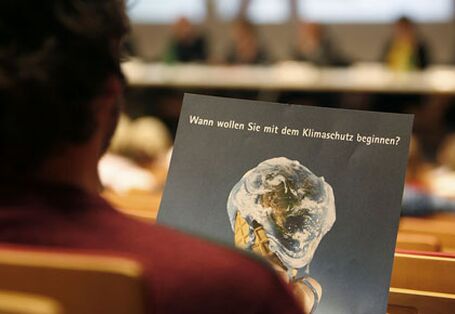 Ein Zuhörer blickt auf eine Broschüre. Das Titelbild zeigt eine Eistüte mit einem verlaufenden Erdball als Weltkugel, darüber steht: "Wann wollen Sie mit dem Klimaschutz beginnen?"