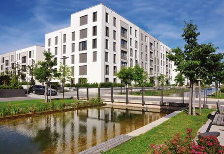 The picture shows an artificial watercourse which runs past a block of flats in a newly developed housing estate. There is a road crossing the watercourse. The watercourse is edged with a narrow strip of vegetation consisting of grass, young trees and flower beds. Benches offer seating. 