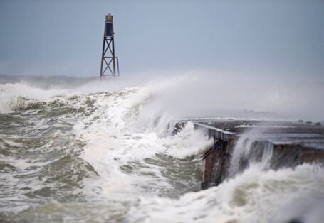 The picture shows a storm surge wave breaking over a coastal building. A metal mast is visible in the background.