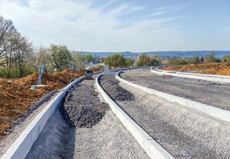 The picture shows the construction of a new road. The partly finished road foundation is visible. In the foreground the foundation is still being completed whereas in the background it is already sealed. Half-way along the site a vibratory plate is standing. To the left of the new road, mounds of earth are visible - behind those a treeline. In the background, the roofs of a residential area are visible.