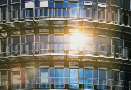 Das Bild zeigt die Glasfront eines Hauses, in der sich die Sonne spiegelt. Einige Fenster sind mit Außenrollos teil-verschattet.